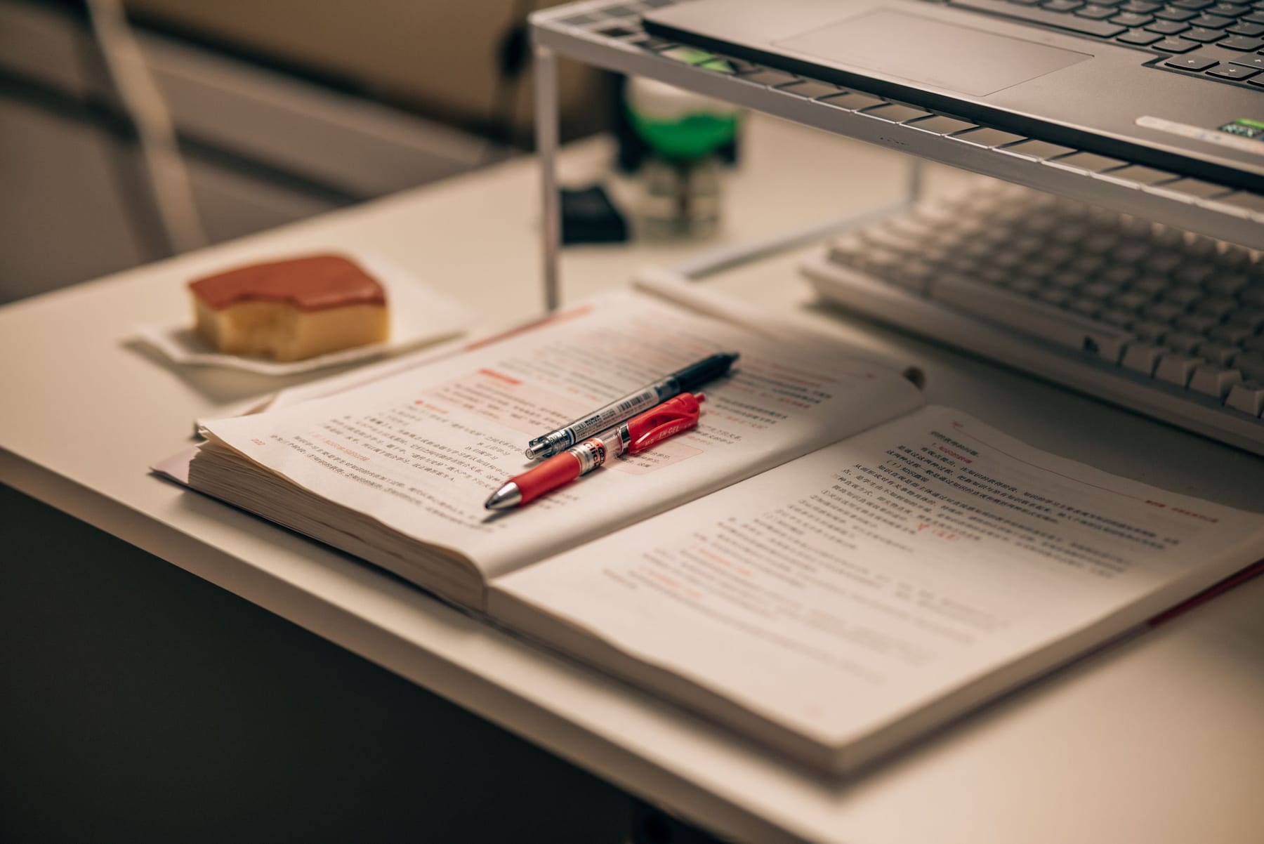 An open textbook with two pens lying on its pages, on a desk beside a keyboard.
