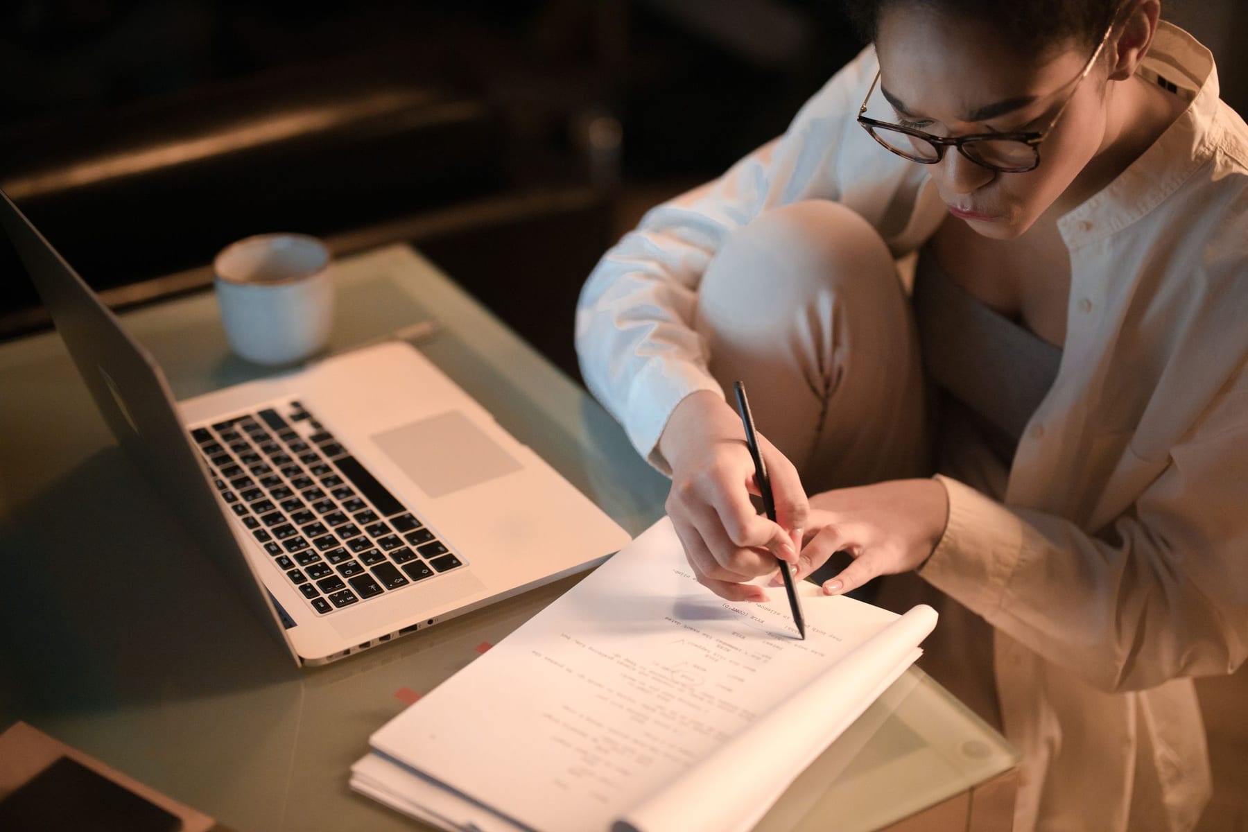 A woman in glasses making notes on a printed page at a low table in the evening, with a laptop open beside her.