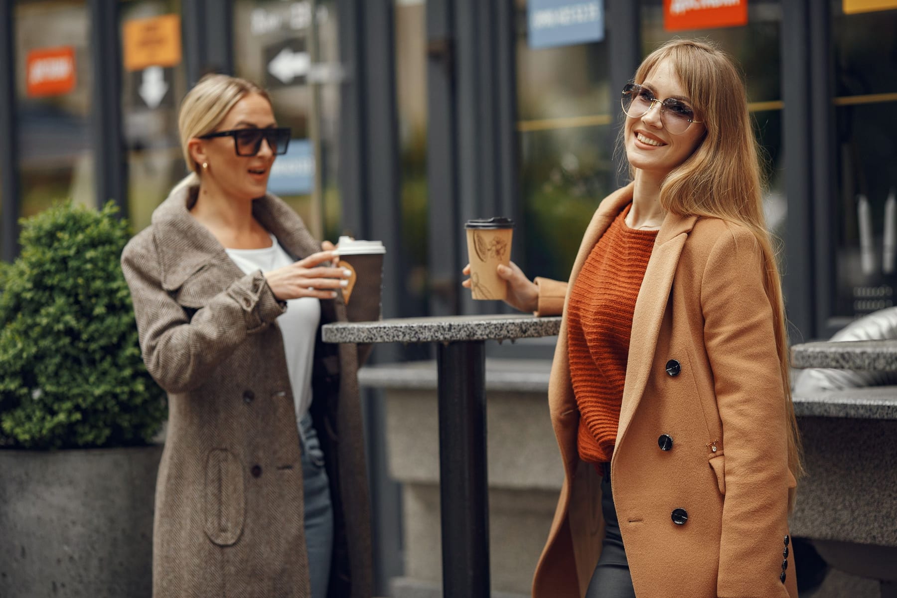 Two women in coats talking over takeaway coffee at a standing table outside a café.
