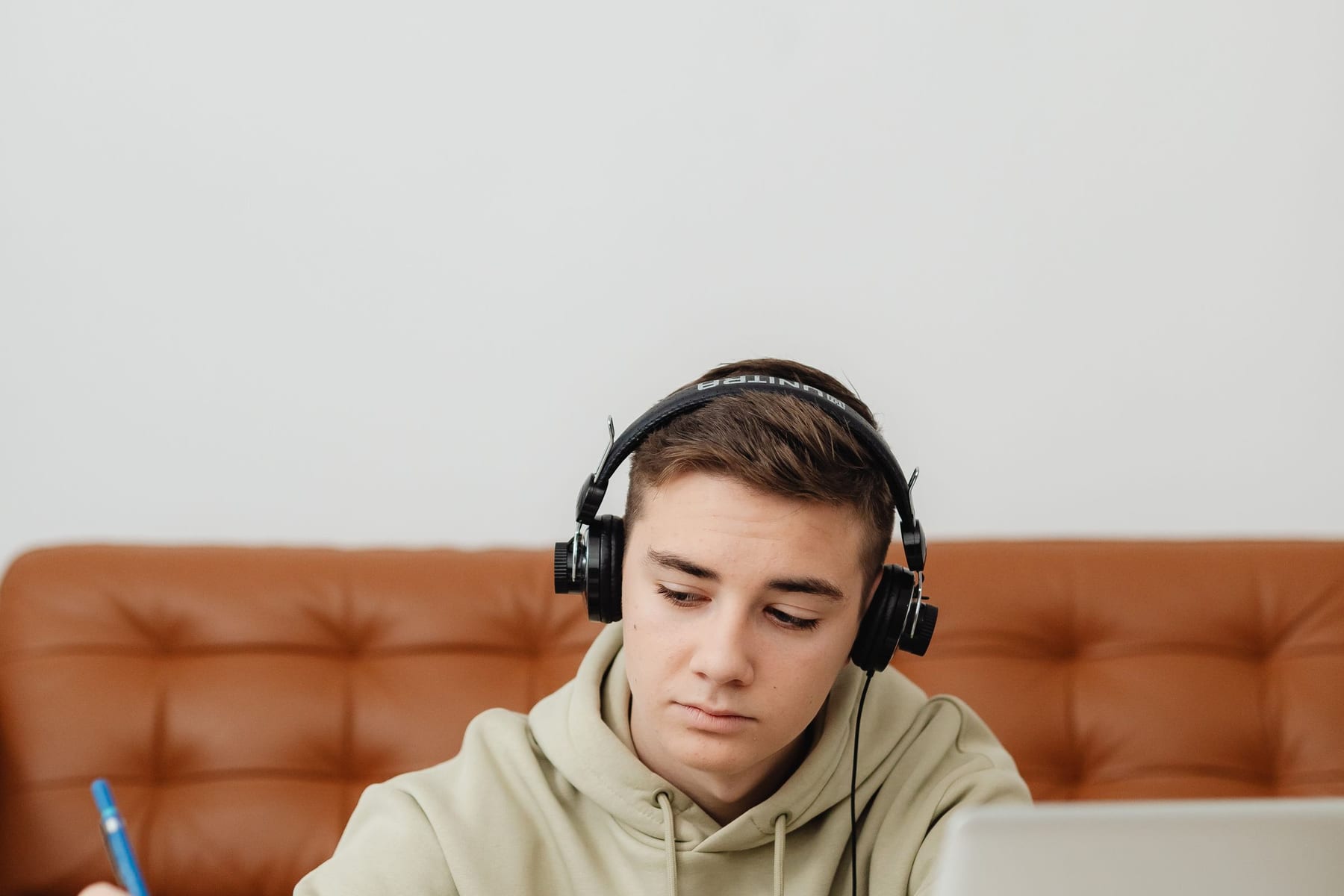 A young man in headphones writing in a notebook beside a laptop.
